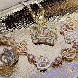 May include: A gold-toned jewelry set featuring a crown pendant with the number 15, a matching ring, and a bracelet with heart-shaped links containing the number 15. The pieces are accented with small clear stones and have a polished metallic finish.