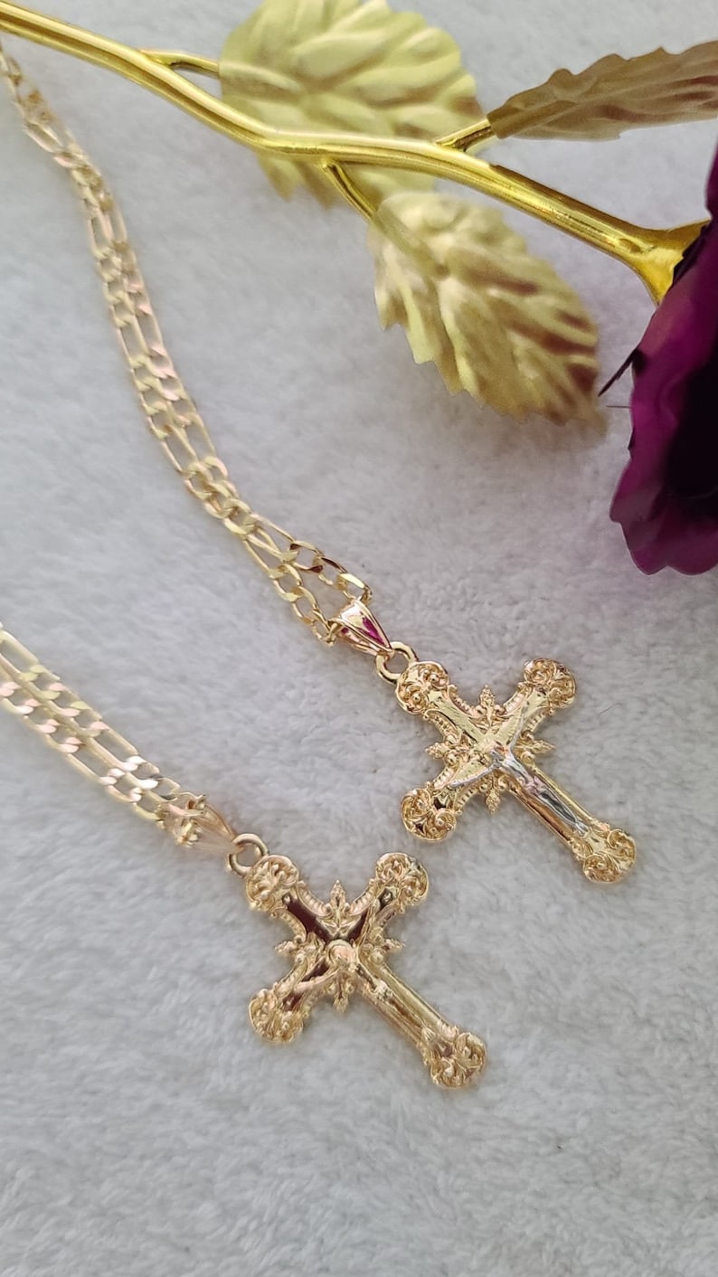 K&ouml;nnte beinhalten: Zwei goldfarbene Kreuzanh&auml;nger mit aufwendigen Details, jeweils an einer passenden Kette befestigt. Die Kreuze weisen komplizierte Designs auf und werden auf einer weichen, wei&szlig;en Oberfl&auml;che mit einer goldfarbenen Rose im Hintergrund pr&auml;sentiert.