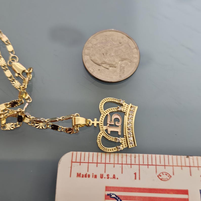 May include: A gold-colored necklace with a crown-shaped pendant featuring the number 15 and small clear stones. A US quarter coin and a ruler showing approximately 1 inch are placed next to the pendant for scale. The chain has a textured link design.