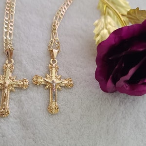 K&ouml;nnte beinhalten: Zwei goldfarbene Kreuzanh&auml;nger mit detaillierten religi&ouml;sen Bildern h&auml;ngen an goldenen Ketten. Im Hintergrund eine lila Rose mit goldfarbenen Bl&auml;ttern. Der Schmuck wird auf einer wei&szlig;en, strukturierten Oberfl&auml;che pr&auml;sentiert.
