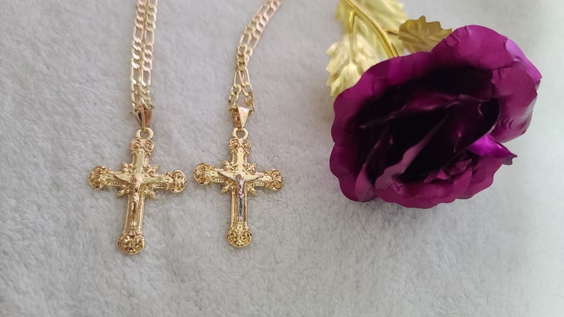 K&ouml;nnte beinhalten: Zwei goldfarbene Kreuzanh&auml;nger mit detaillierten religi&ouml;sen Bildern h&auml;ngen an goldenen Ketten. Im Hintergrund eine lila Rose mit goldfarbenen Bl&auml;ttern. Der Schmuck wird auf einer wei&szlig;en, strukturierten Oberfl&auml;che pr&auml;sentiert.