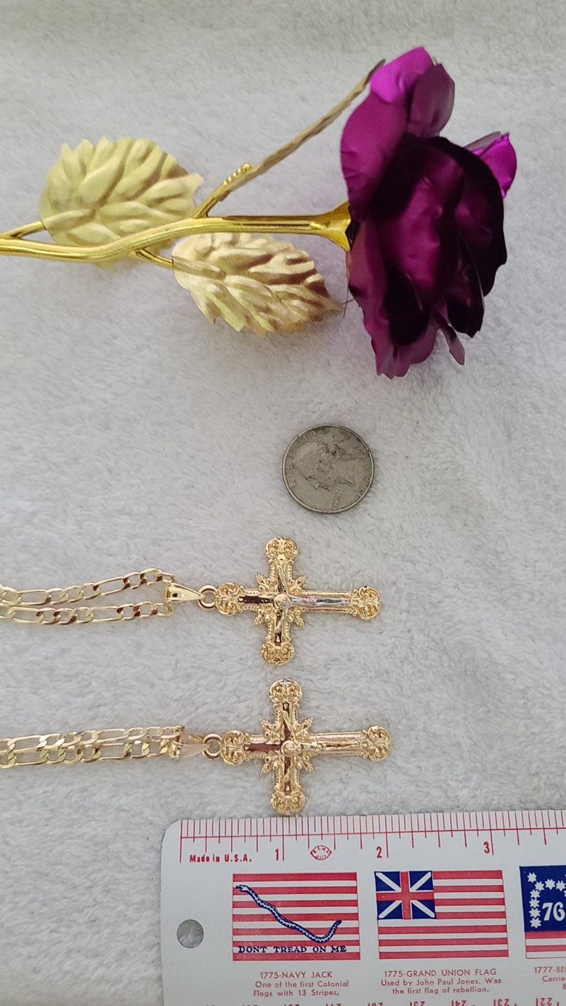 K&ouml;nnte beinhalten: Zwei goldfarbene Kreuzanh&auml;nger mit aufwendigen Details, jeweils an einer passenden Kette. Eine lila Rose mit goldfarbenen Bl&auml;ttern und eine US-Vierteldollarm&uuml;nze sind ebenfalls vorhanden. Ein Lineal mit dem Text "Made in U.S.A." ist sichtbar.