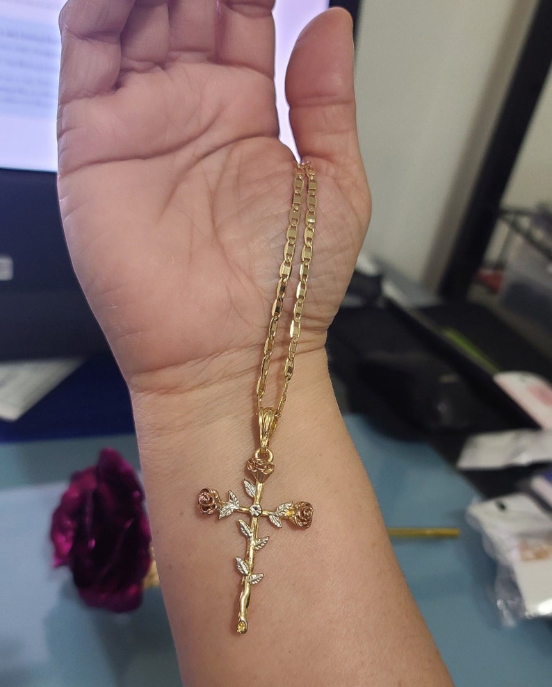 May include: A gold chain necklace with a rose cross pendant. The cross is made of gold and has three roses on it. The roses are a mix of gold and white.