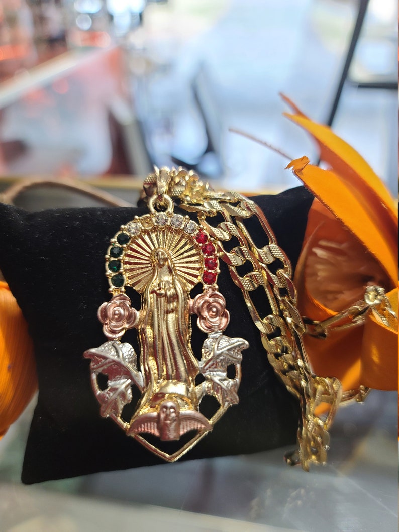 May include: Gold pendant necklace featuring a detailed depiction of the Virgin Mary with red, green, and white gemstones. The pendant is surrounded by gold leaves and a dove.