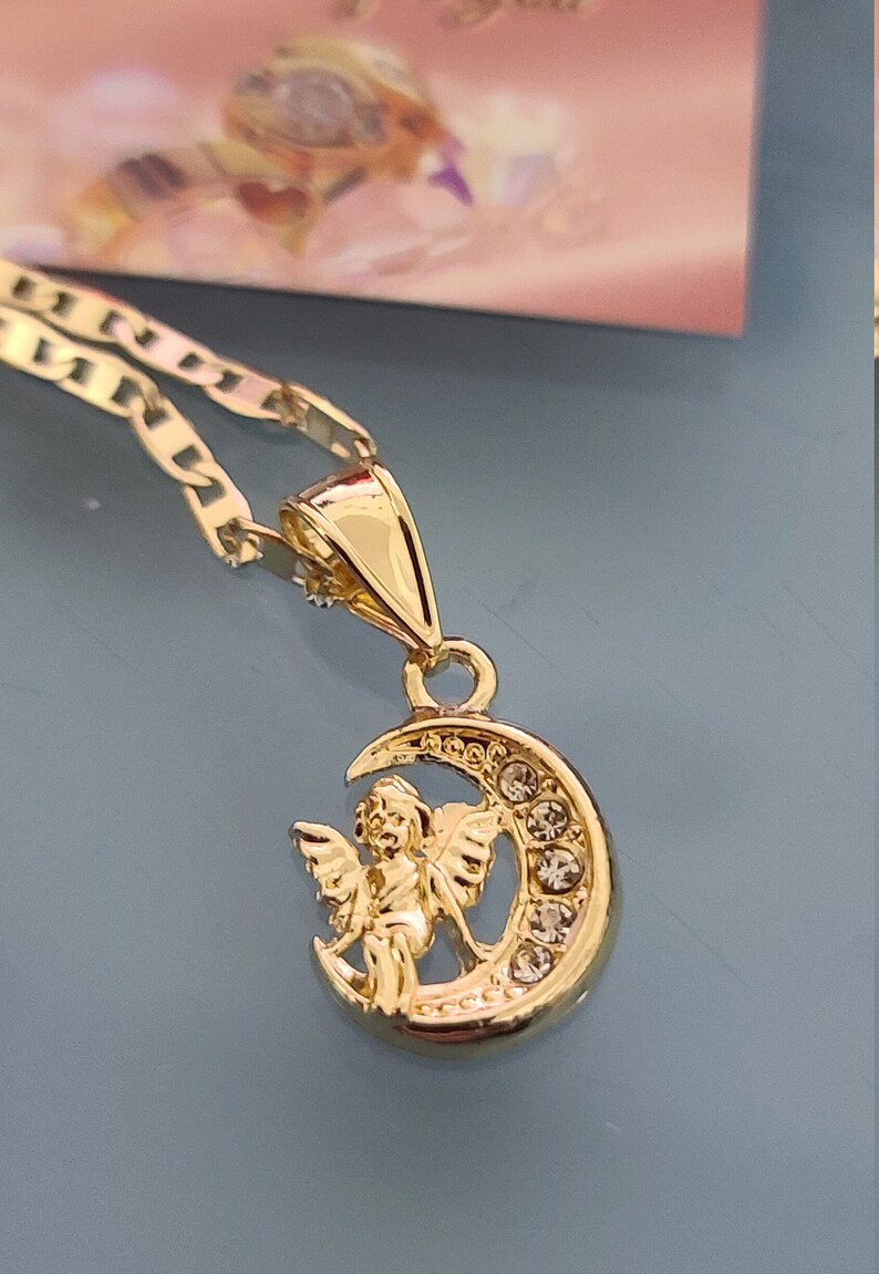 May include: Gold necklace with a pendant featuring a cherub sitting on a crescent moon with clear crystals.