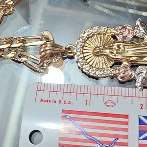 May include: A gold chain necklace with a pendant featuring a three-dimensional depiction of the Virgin Mary. The pendant is made of gold, silver, and rose gold and features a halo, roses, and leaves. The chain is a curb chain style. The pendant is approximately 2 inches in length. "Made in U.S.A." is printed on a ruler in the background.