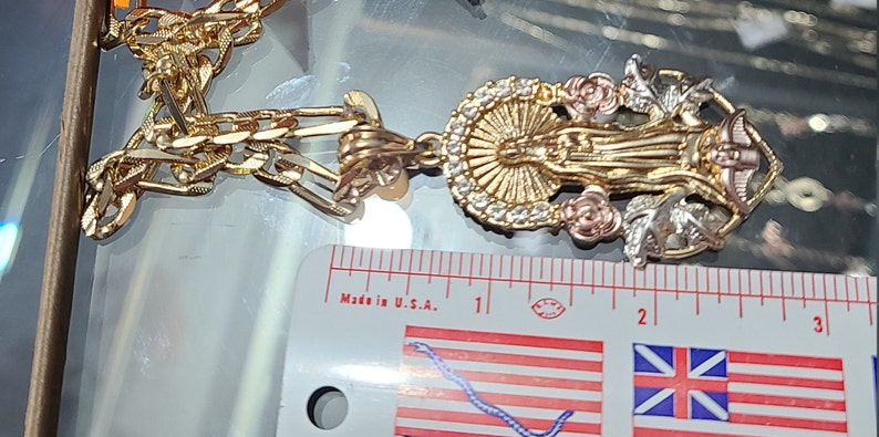 May include: A gold chain necklace with a pendant featuring a three-dimensional depiction of the Virgin Mary. The pendant is made of gold, silver, and rose gold and features a halo, roses, and leaves. The chain is a curb chain style. The pendant is approximately 2 inches in length. "Made in U.S.A." is printed on a ruler in the background.