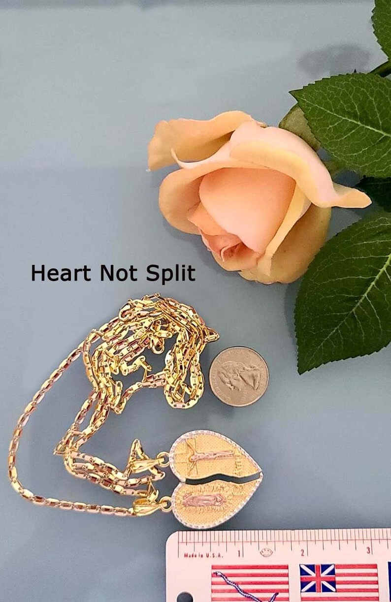 May include: A gold chain necklace with a heart-shaped pendant featuring a religious image. The pendant is split in two, with each half depicting a different figure. The chain is made of multiple strands of gold links. The necklace is displayed on a blue surface with a rose and a ruler for scale.