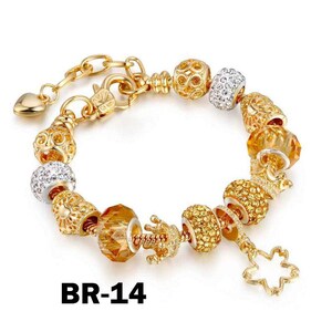 May include: Gold-toned charm bracelet with various decorative beads, including crystal-studded spheres, faceted amber-colored beads, and a crown charm. The bracelet features a heart-shaped clasp and a dangling flower charm. The text "BR-14" is visible.
