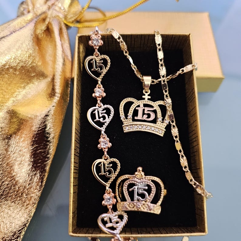May include: A gold-toned jewelry set featuring a necklace with a crown pendant, a bracelet with heart-shaped links, and a crown ring. Each piece displays the number 15 and is accented with small clear stones. The set is presented in a gold box.