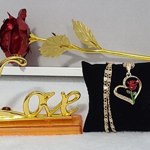 May include: A gold-plated rose with a red velvet rose head, a gold 'Love' sign, and a gold necklace with a heart-shaped pendant featuring a red rose. The necklace is displayed on a black velvet cushion.