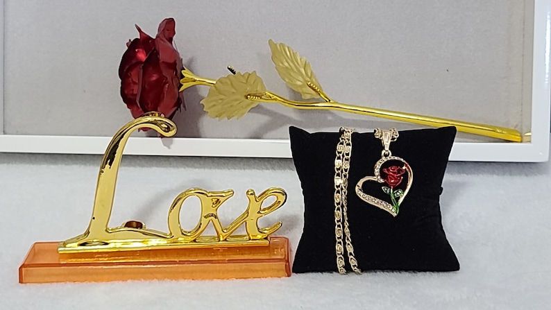 May include: A gold-plated rose with a red velvet rose head, a gold 'Love' sign, and a gold necklace with a heart-shaped pendant featuring a red rose. The necklace is displayed on a black velvet cushion.
