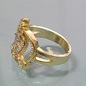 May include: A gold-toned ring featuring a split shank design that leads to a central, elevated rectangular setting. The setting is framed by a textured border and contains a clear, faceted stone that reflects light, creating a bright appearance.