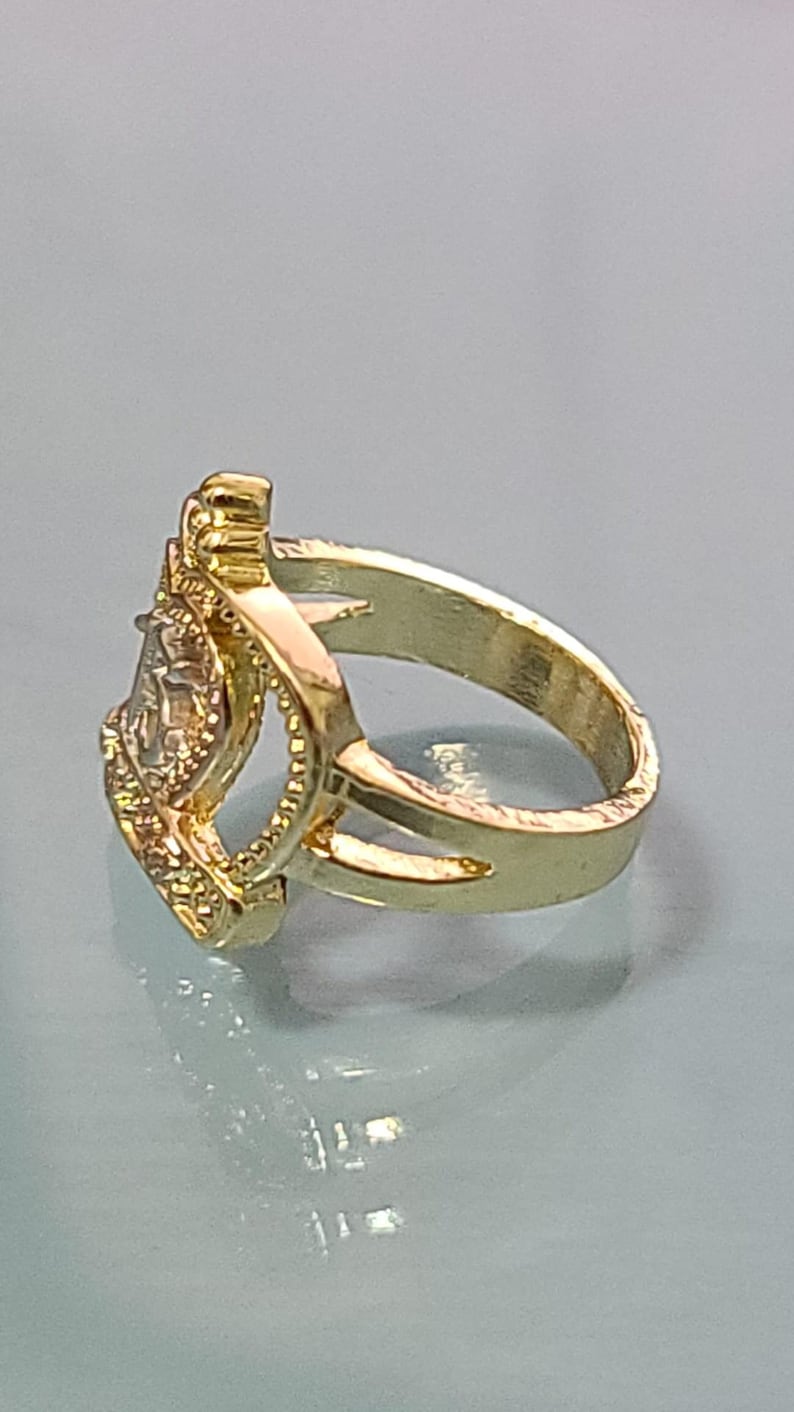 May include: A gold-toned ring featuring a split shank design that leads to a central, elevated rectangular setting. The setting is framed by a textured border and contains a clear, faceted stone that reflects light, creating a bright appearance.