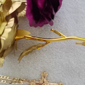 K&ouml;nnte beinhalten: Ein goldfarbener Kreuzanh&auml;nger mit einem verzierten Design, der auf einer hellgrauen Oberfl&auml;che liegt. Eine goldfarbene Rose und eine lila Rose sind ebenfalls im Bild, was auf ein religi&ouml;ses oder dekoratives Thema hindeutet.