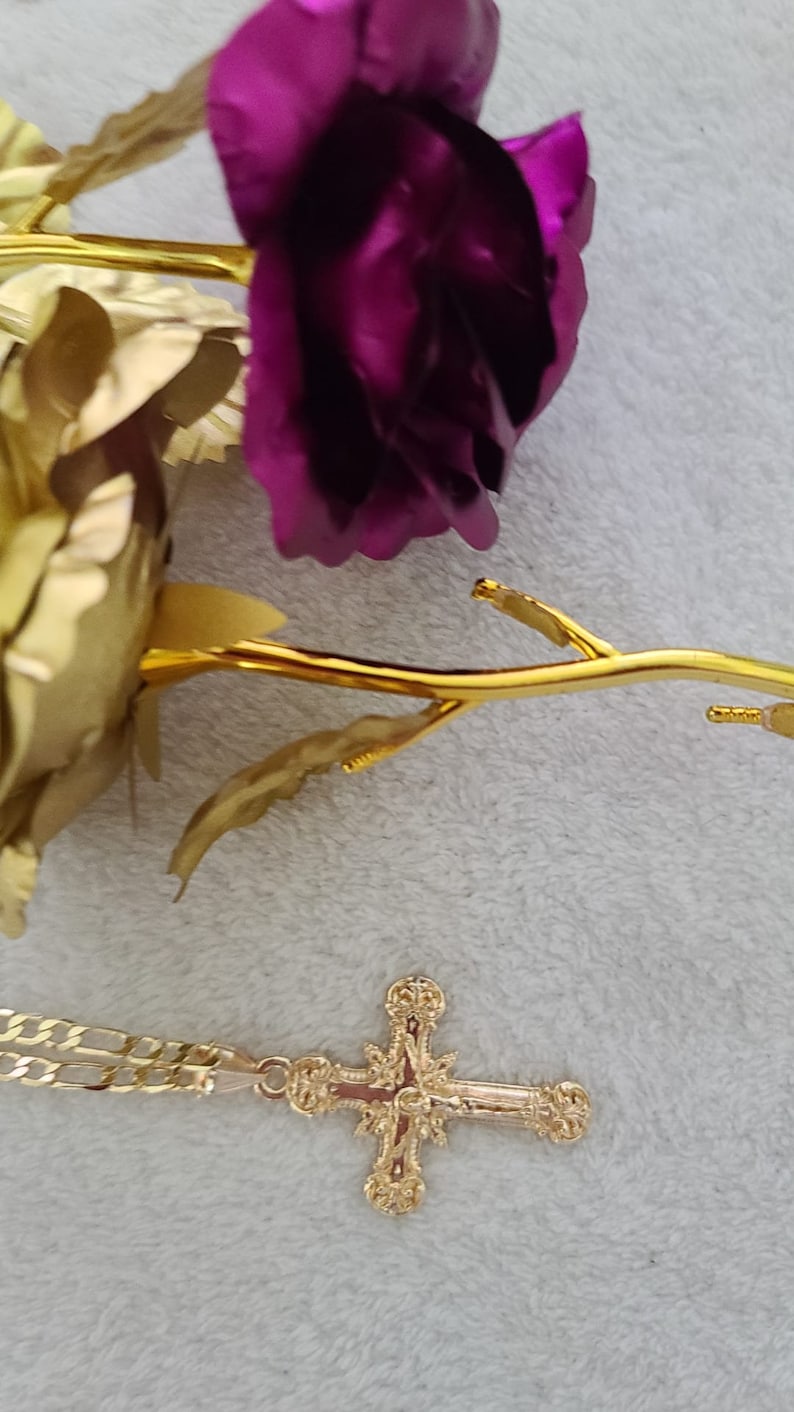 K&ouml;nnte beinhalten: Ein goldfarbener Kreuzanh&auml;nger mit einem verzierten Design, der auf einer hellgrauen Oberfl&auml;che liegt. Eine goldfarbene Rose und eine lila Rose sind ebenfalls im Bild, was auf ein religi&ouml;ses oder dekoratives Thema hindeutet.