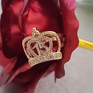 May include: A gold-toned ring featuring a crown-shaped centerpiece with the number 15 prominently displayed in the center. The crown design is accented with small, clear, sparkling stones along the base and the upper arched frame.