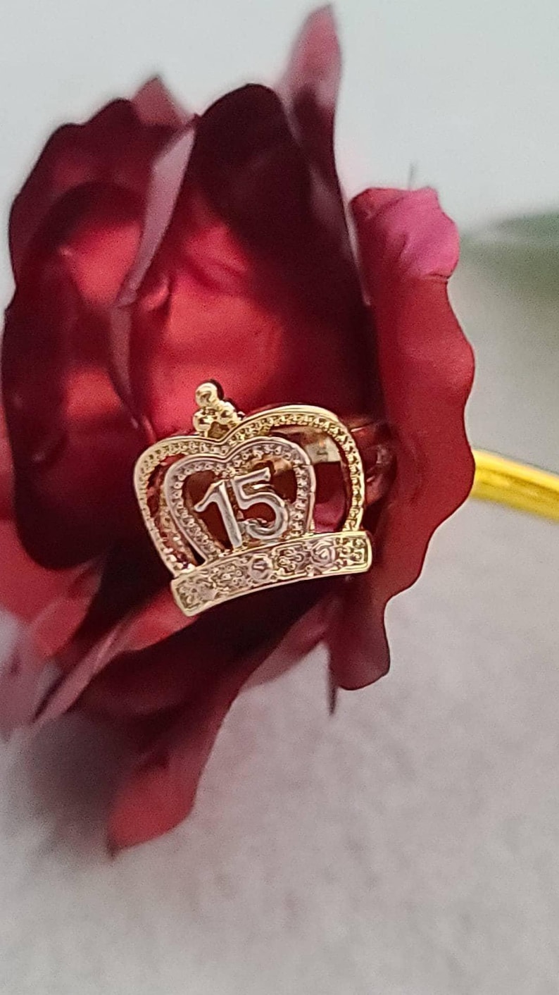 May include: A gold-toned ring featuring a crown-shaped centerpiece with the number 15 prominently displayed in the center. The crown design is accented with small, clear, sparkling stones along the base and the upper arched frame.