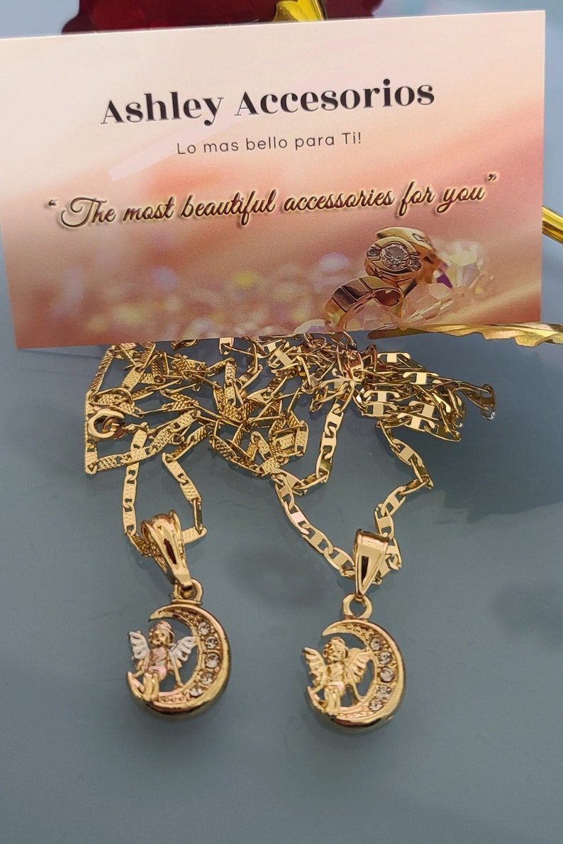 May include: Two gold necklaces with angel and crescent moon pendants. The necklaces are on a blue background. The text "Ashley Accesorios" and "Lo mas bello para Ti!" is visible on a card behind the necklaces.
