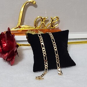 K&ouml;nnte beinhalten: Eine goldfarbene Figaro-Kettenhalskette auf einem schwarzen Samtkissen. Eine rote Metallrose und ein goldenes "Love"-Zeichen befinden sich im Hintergrund. Die Halskette hat ein sich wiederholendes Muster aus drei kleinen Gliedern, gefolgt von einem l&auml;nglichen Glied.