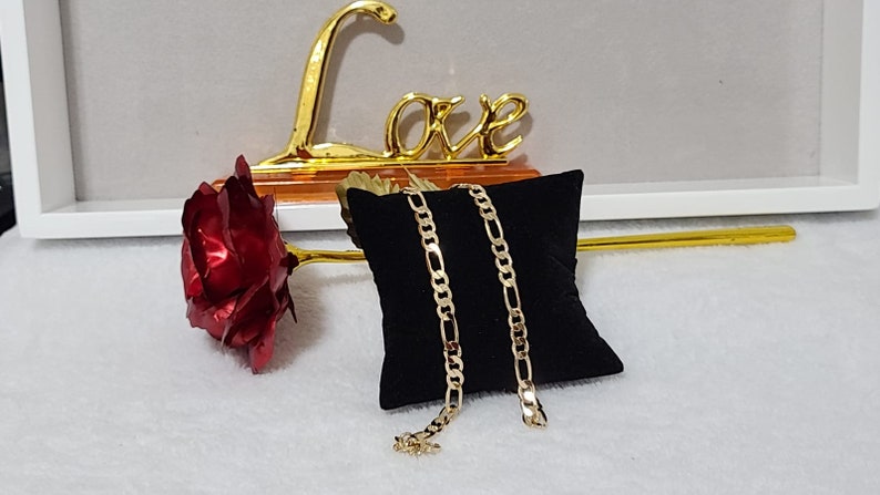 K&ouml;nnte beinhalten: Eine goldfarbene Figaro-Kettenhalskette auf einem schwarzen Samtkissen. Eine rote Metallrose und ein goldenes "Love"-Zeichen befinden sich im Hintergrund. Die Halskette hat ein sich wiederholendes Muster aus drei kleinen Gliedern, gefolgt von einem l&auml;nglichen Glied.