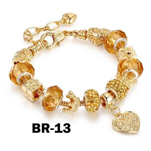 May include: Gold-toned charm bracelet with various decorative beads, including faceted amber-colored glass beads, a crown, and a heart charm. The bracelet has a lobster clasp closure and a heart-shaped charm. The text "BR-13" is printed on the bottom.