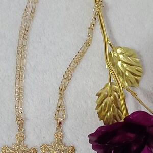 K&ouml;nnte beinhalten: Zwei goldfarbene Kreuzketten mit aufwendigen Details h&auml;ngen auf einer wei&szlig;en Oberfl&auml;che. Die Kreuze sind an filigranen Ketten aufgeh&auml;ngt. Ein goldfarbener Rosenstiel mit Bl&auml;ttern und eine tiefviolette Rosenbl&uuml;te befinden sich im Hintergrund.