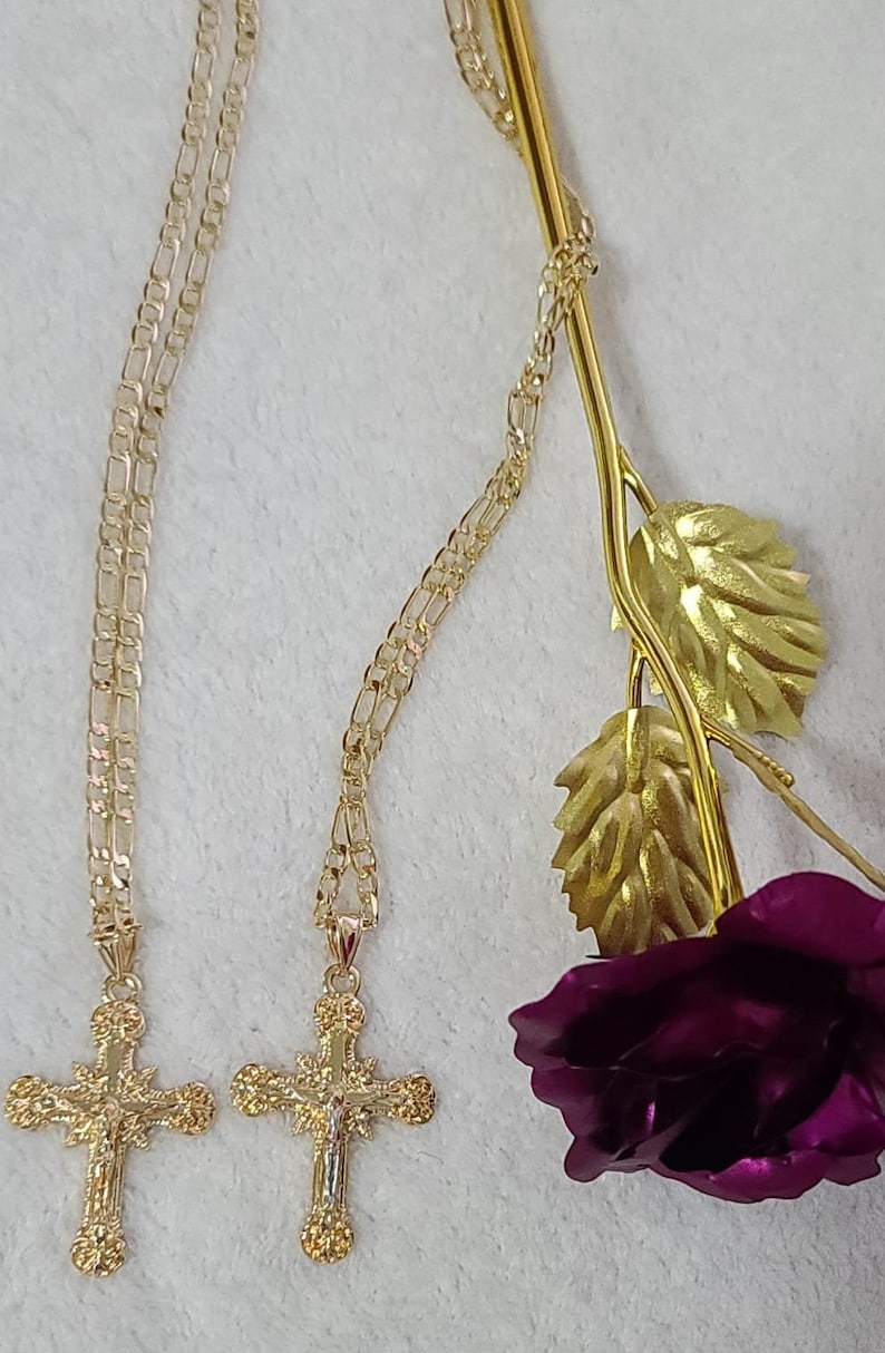 K&ouml;nnte beinhalten: Zwei goldfarbene Kreuzketten mit aufwendigen Details h&auml;ngen auf einer wei&szlig;en Oberfl&auml;che. Die Kreuze sind an filigranen Ketten aufgeh&auml;ngt. Ein goldfarbener Rosenstiel mit Bl&auml;ttern und eine tiefviolette Rosenbl&uuml;te befinden sich im Hintergrund.