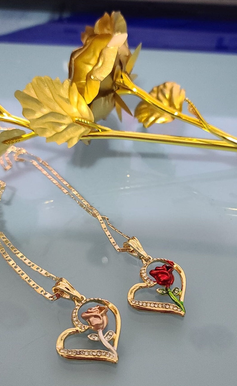 May include: Two gold necklaces with heart-shaped pendants featuring a rose design. One pendant has a red rose, the other has a pink rose.