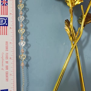 May include: A gold-tone bracelet featuring a series of heart-shaped links, each containing the number 15, alternating with small clear stones. A ruler placed alongside shows the bracelet measures approximately 7.5 inches in total length.