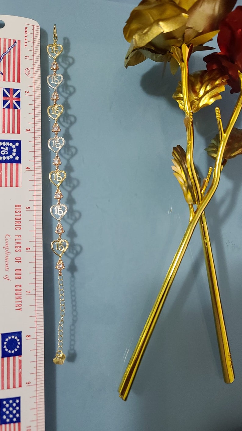 May include: A gold-tone bracelet featuring a series of heart-shaped links, each containing the number 15, alternating with small clear stones. A ruler placed alongside shows the bracelet measures approximately 7.5 inches in total length.