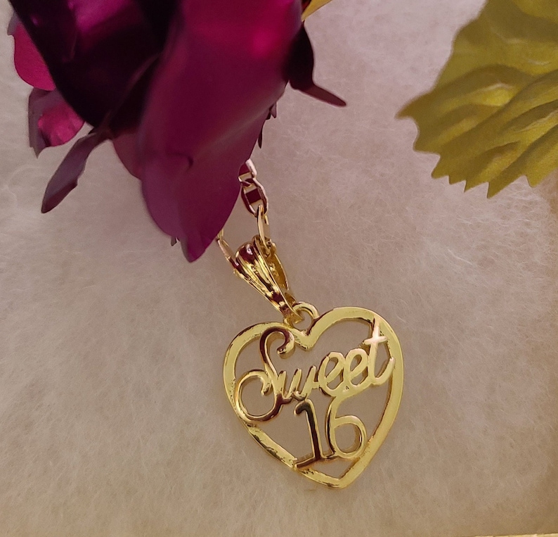 May include: Gold heart-shaped pendant necklace with the words "Sweet 16" inscribed inside. The necklace is hanging from a rose.