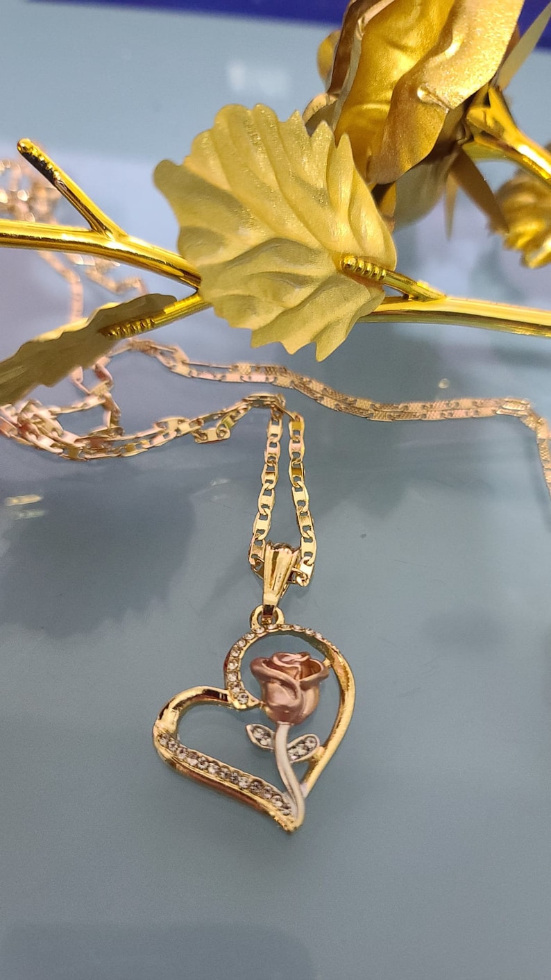 May include: A gold heart-shaped pendant necklace with a rose design. The rose is pink and white and has a stem with leaves. The necklace has a delicate chain.