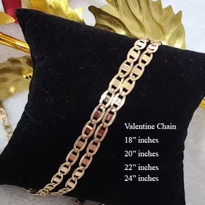 May include: A gold-colored chain necklace on a black velvet cushion. The chain is a mariner style with oval links. The text "Valentine Chain" is printed on the cushion, along with the available sizes: 18", 20", 22", and 24" inches.