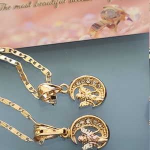 May include: Two gold necklaces with angel and crescent moon pendants. The pendants are adorned with clear rhinestones. The necklaces are on a blue surface with a ruler that reads "Made in U.S.A."