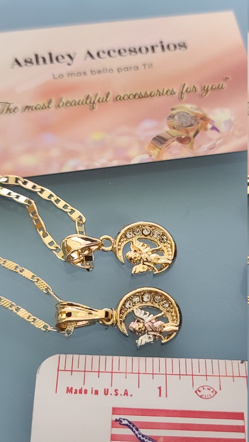 May include: Two gold necklaces with angel and crescent moon pendants. The pendants are adorned with clear rhinestones. The necklaces are on a blue surface with a ruler that reads "Made in U.S.A."
