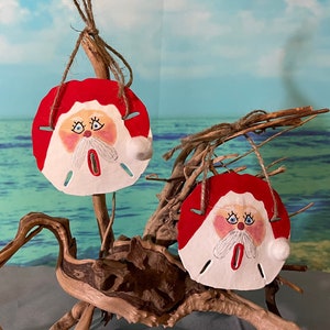 May include: Two hand-painted sand dollar ornaments with Santa Claus faces. The ornaments are red, white, and blue and are hanging from a piece of driftwood.