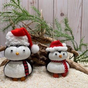 May include: Two Christmas penguin ornaments. One penguin wears a red and white Santa hat and a black and red plaid scarf. The other penguin wears a red and white Santa hat and a red and white scarf. Both have orange feet and beaks.