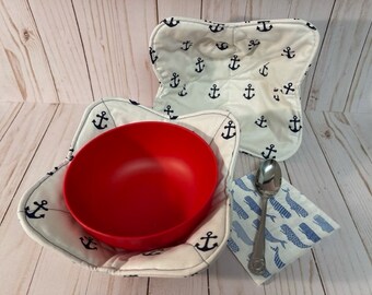 May include: Two white fabric bowl cozies with a navy blue anchor pattern. A red bowl is inside one of the cozies. A blue and white fabric napkin with a whale pattern is on a wooden surface with a silver spoon on top.