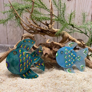 May include: Two hand-painted wooden fish ornaments. The larger fish is teal blue with yellow and purple accents. The smaller fish is light blue with green and white accents.