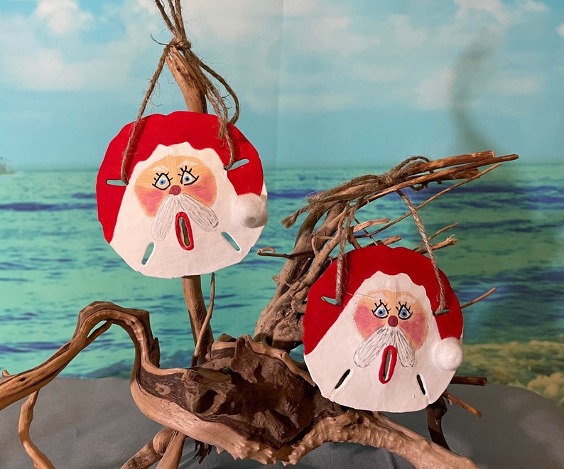 May include: Two hand-painted sand dollar ornaments with Santa Claus faces. The ornaments are red, white, and blue and are hanging from a piece of driftwood.