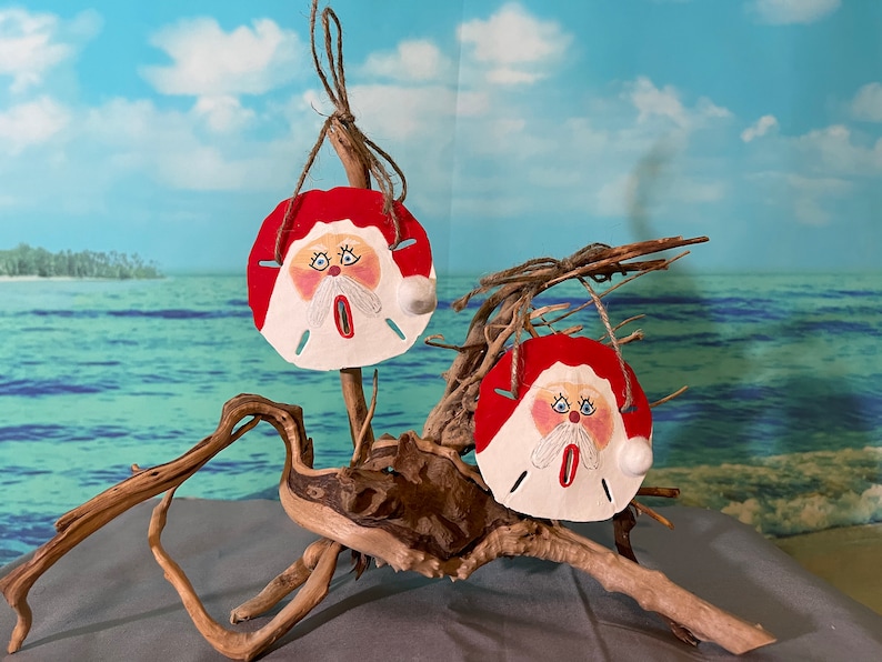 May include: Two painted sand dollar ornaments with Santa Claus faces, hanging from driftwood against a blue sky and ocean background.