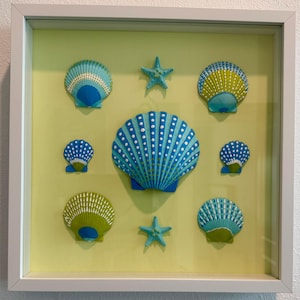 May include: A square white shadow box frame containing a collection of blue, green, and white seashells and two starfish arranged on a pale yellow background. The central shell is the largest, surrounded by six smaller shells and two starfish.