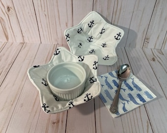 May include: Two white star-shaped fabric pot holders with a navy blue anchor print. A white ceramic bowl sits inside one of the pot holders. A white napkin with a blue whale print and a silver spoon are on the table.