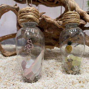 May include: Two glass ornaments with a rope and twine top. One ornament contains a pink flamingo charm and sea glass, the other contains a pineapple charm and sea glass.