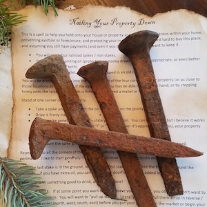 May include: A close-up of four rusty railroad spikes on a piece of parchment paper with text that reads "Nailing Your Property Down". The text describes a spell to help hold onto your house or property and protect it from eviction or foreclosure.