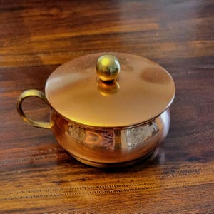 May include: A polished copper bowl with a matching lid and a single side handle. The lid features a small, spherical brass-colored knob at the center. The metallic surface is reflective, showing a warm, reddish-brown tone throughout the piece.
