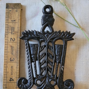 May include: Black cast iron trivet with a design of three brooms and leaves. The trivet has a loop at the top for hanging.