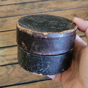 May include: A round, black, lidded container with a textured surface and visible signs of wear along the edges. The cylindrical box features a dark, weathered finish with brown undertones showing through the worn areas of the lid and base rim.
