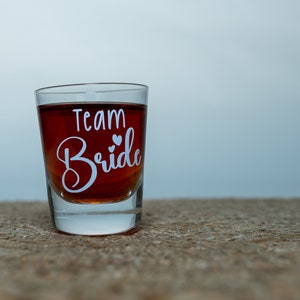 Op de afbeelding: Een helder shotglaasje gevuld met een donkere vloeistof. Het glas heeft de tekst "Team Bride" erop geschreven in wit.
